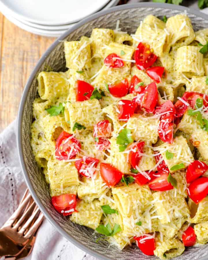 overhead shot of creamy pesto pasta in a grey bowl with diced tomatoes on top on a wood surface