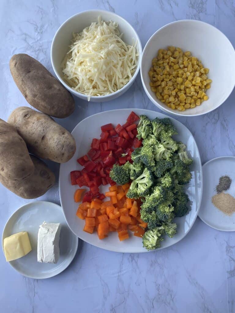 overhead shot of ingredients on a white marble surface. 4 Idaho potatoes, butter, cream cheese, bell peppers, broccoli, white Cheddar, corn, garlic powder, salt and papper.