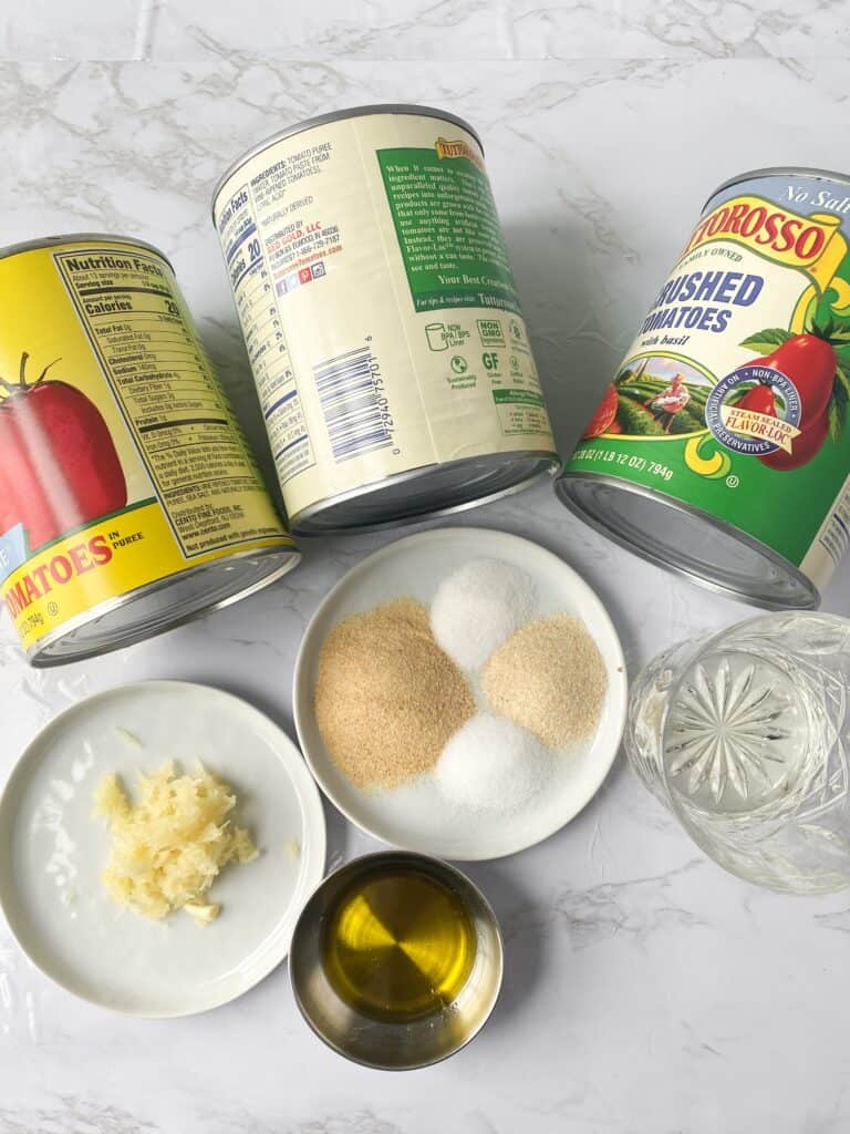 overhead photo of the ingredients on a marble surface: two cans of tomato puree, one can of crushed tomatoes, white wine, garlic, extra virgin olive oil, garlic powder, onion powder, salt, pepper, and sugar.