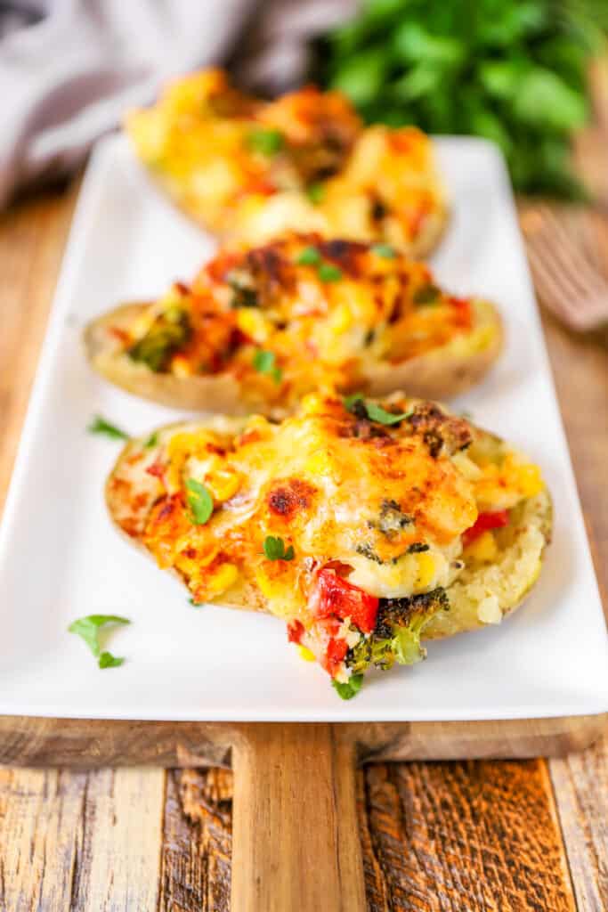 the veggie stuffed potatoes on a white dish on a wood surface. Parsley, grey napkin, and forks in the background.