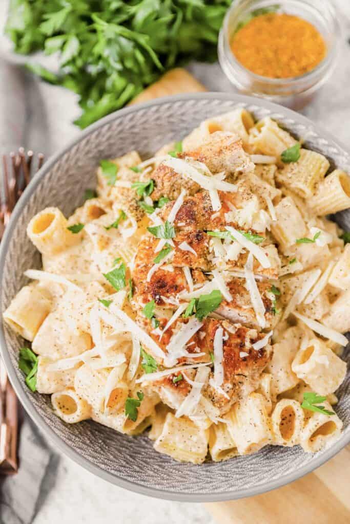 Cajun seasoned chicken on top of pasta in a grey bowl. Parsley and jar of spices in the back.