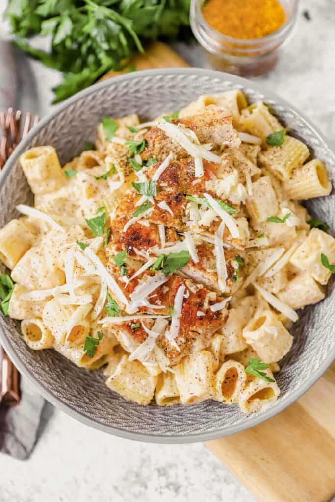 Cajun seasoned chicken on top of pasta in a grey bowl on a wood board. Parsley and jar of spices in the back.