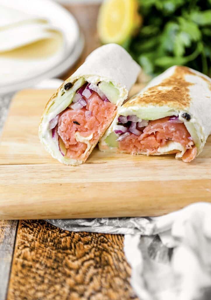 the two pieces of the smoked salmon wrap on a wood board. In the background some parsley, tortillas, and a lemon wedge.