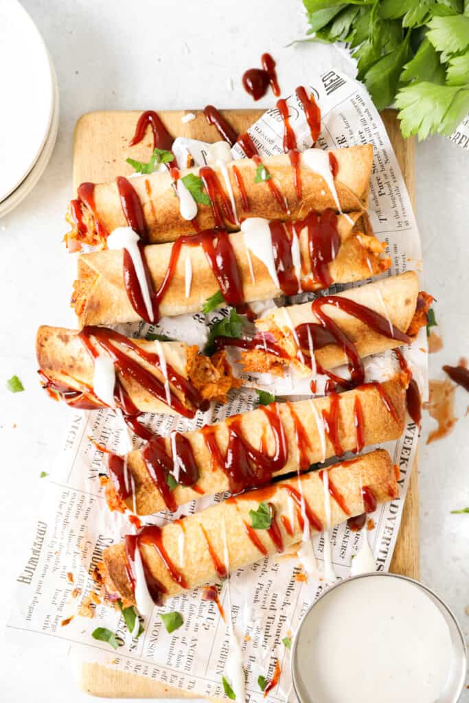 overhead shot of bbq chicken taquitos drizzled with ranch and bbq sauce on white newsprint paper.