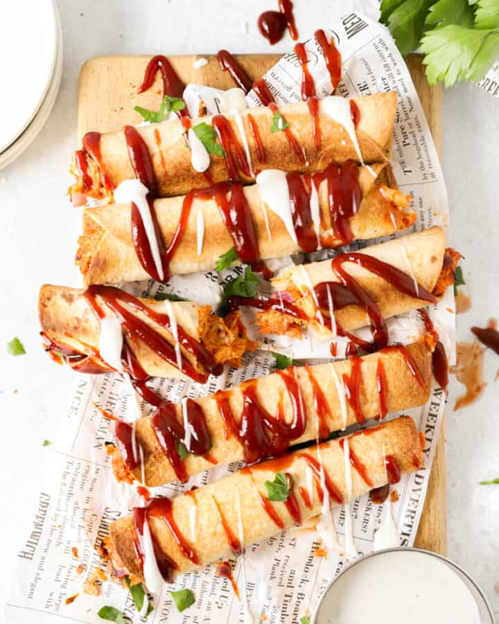 overhead shot of bbq chicken taquitos drizzled with ranch and bbq sauce on white newsprint paper.