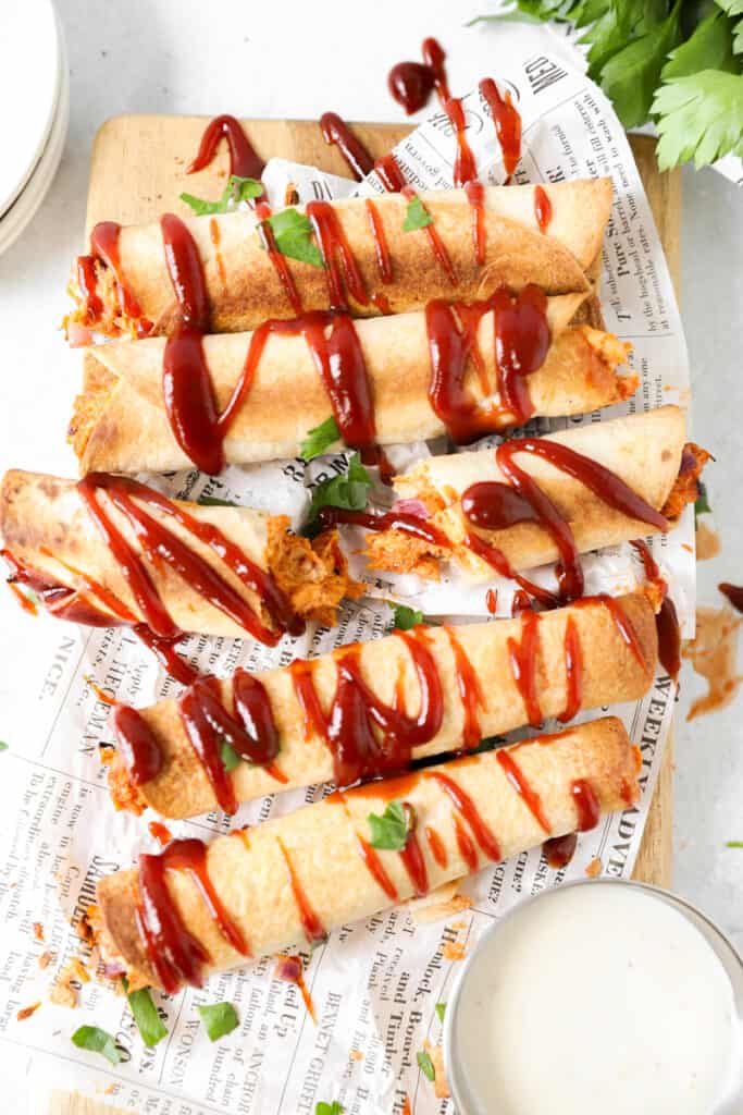 overhead shot of bbq chicken taquitos drizzled with ranch and bbq sauce on white newsprint paper. Ranch and parsley off to the side.