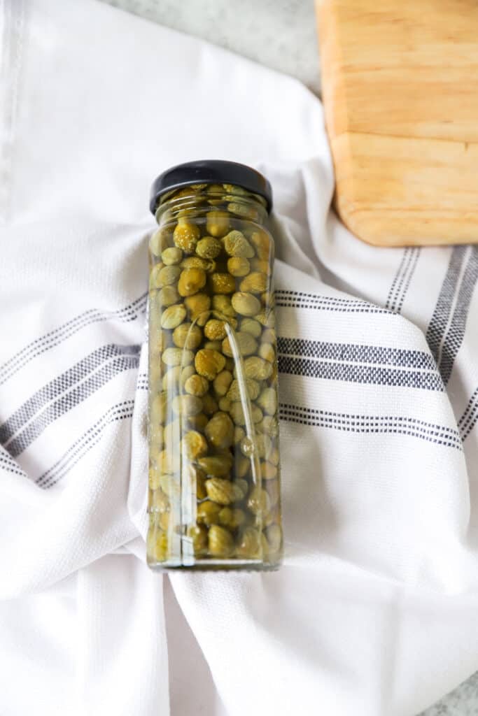 jar of capers on a blue striped white linen and a wood cutting board off to the side.