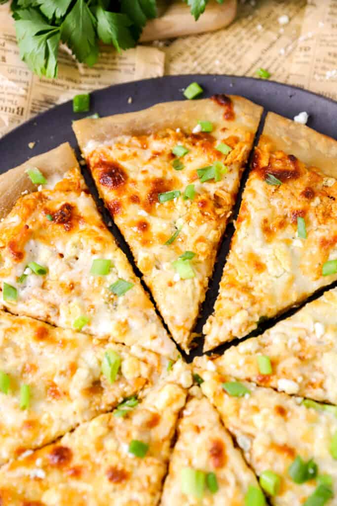 Buffalo chicken slices with diced scallions on a pizza pan with parsley in the back.