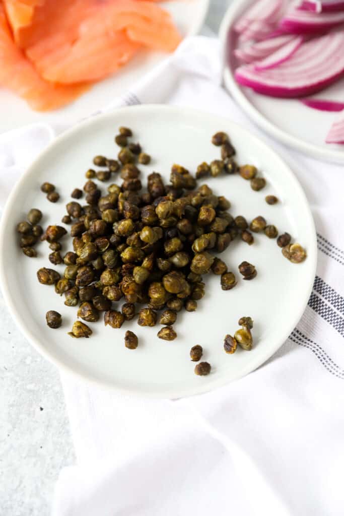 fried capers on a small white plate, in the background smoked salmon and red onions