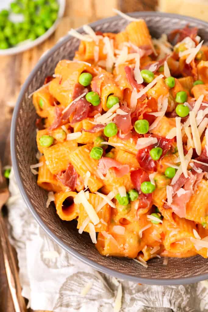 side view of the large bowl of pasta on a wood surface with a plate of peas in the back.