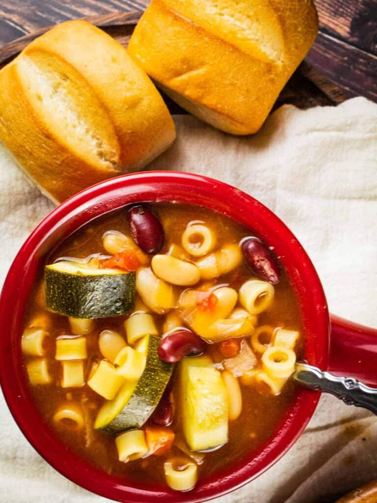 overhead shot of the minestrone soup in a red cup with two bread rolls.