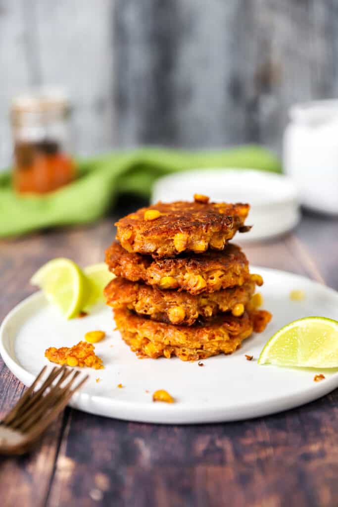 stack of four fritters on a white plate with lime wedges on a wood surface.