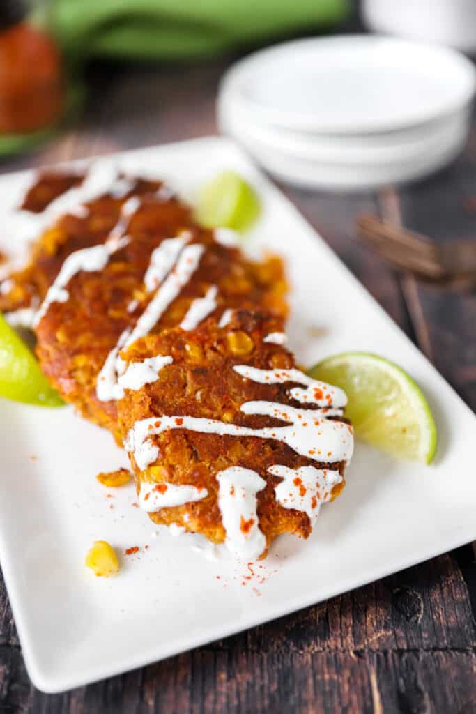 four spicy corn fritters layered on a rectangle white platter with lime wedges on a wood surface.