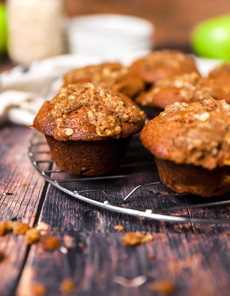 apple muffins on a baking rack on a wood surface, plates and linen in the back.
