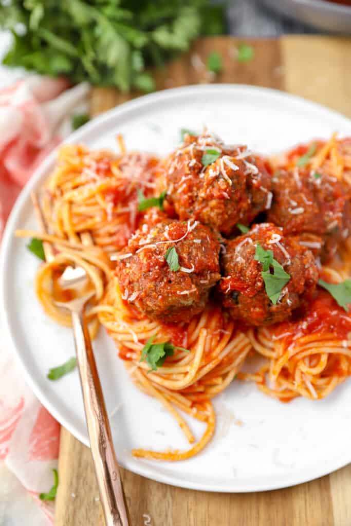 a white plate with spiraled thin spaghetti, a bronze fork, and several meatballs topped with Parmesan and parsley.
