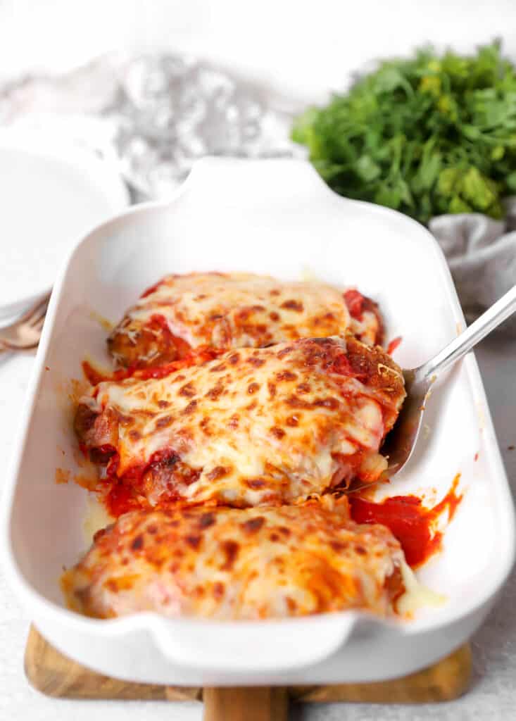 a stainless steel spatula scooping once of the chicken parms from a white casserole dish.