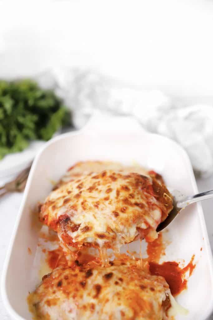 scooping the roasted red pepper chicken parm from the white baking dish with a stainless steel spatula.