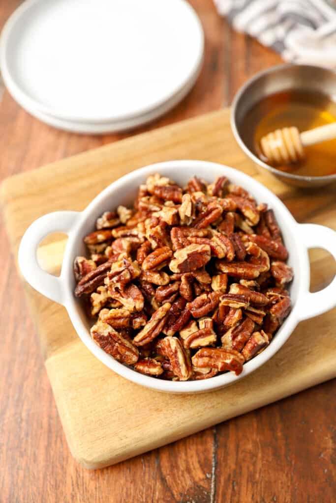 a white dish with the honey roasted pecans on a wood cutting board