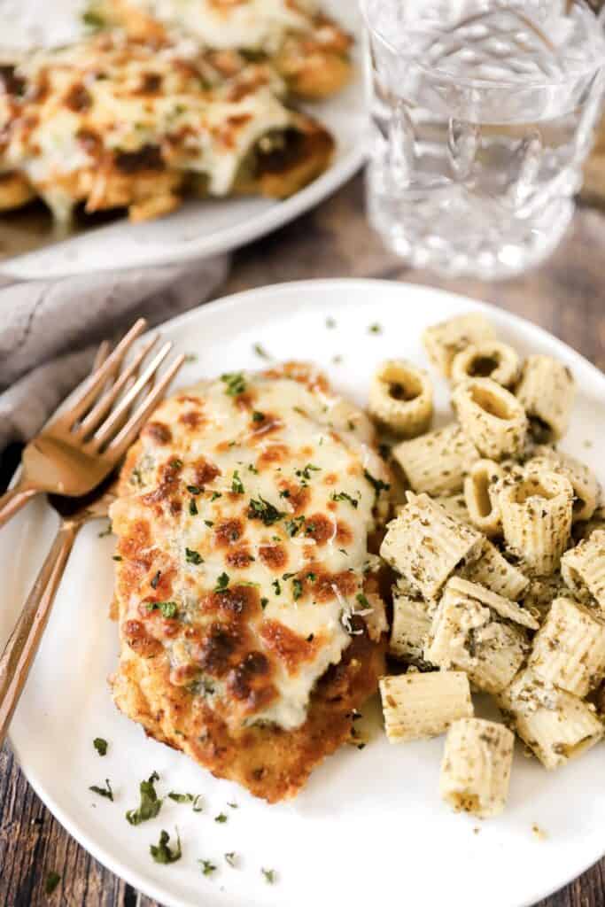 a plate with one pesto chicken parm, some rigatoni, an two  rose gold forks.