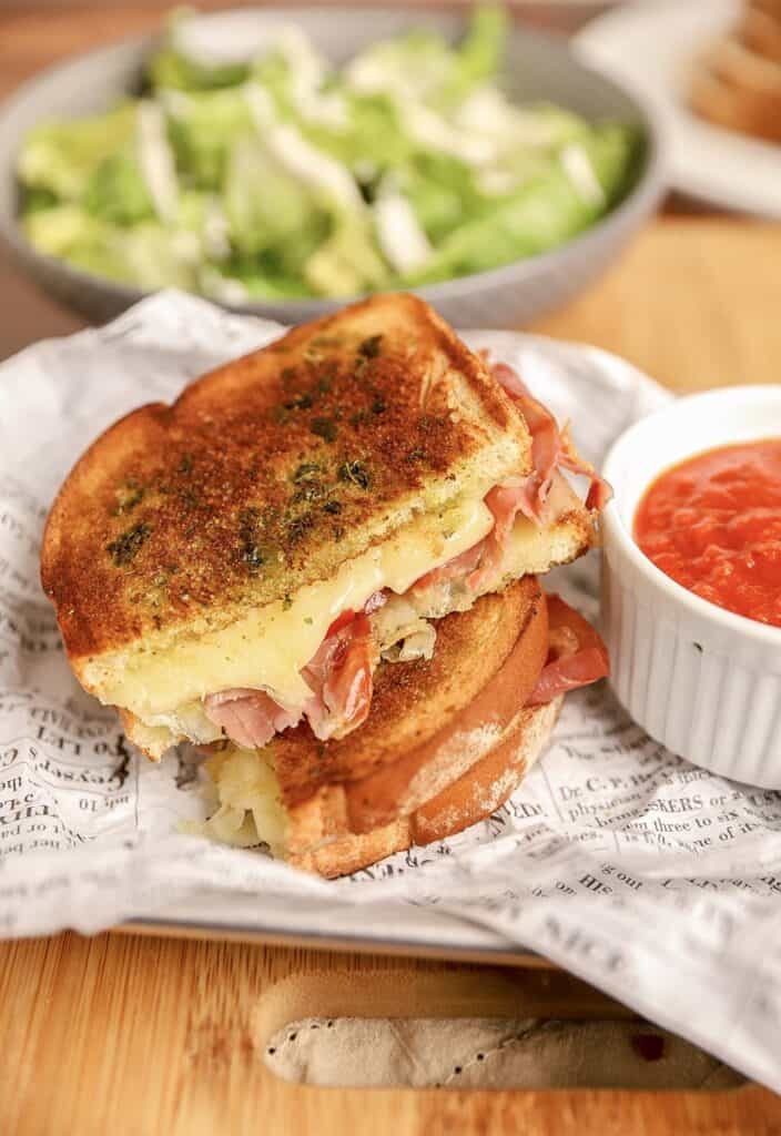 an Italian grilled cheese on newsprint paper with a white ramekin of marinara sauce.