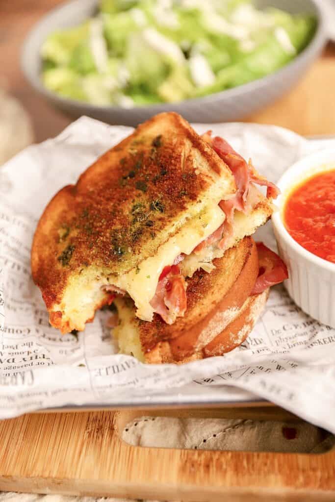 an Italian grilled cheese on newsprint paper with a white ramekin of marinara sauce.