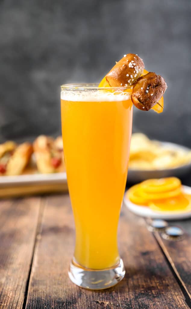 a beermosa on a wood surface with oranges and hotdog with chips in the background.