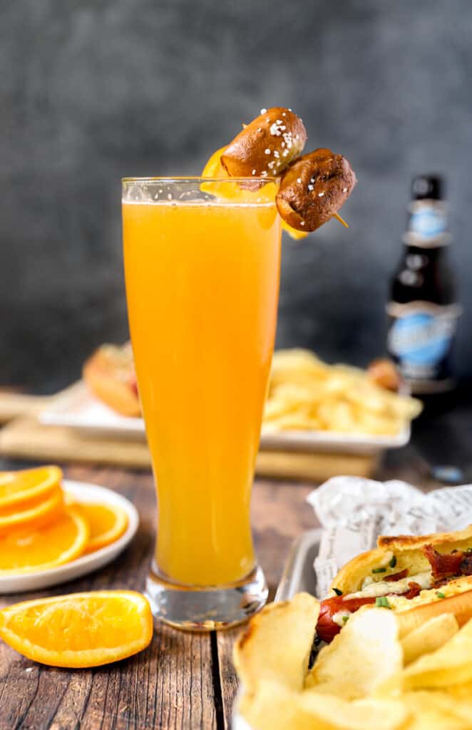 a beermosa on a wood surface with oranges and hotdog with chips