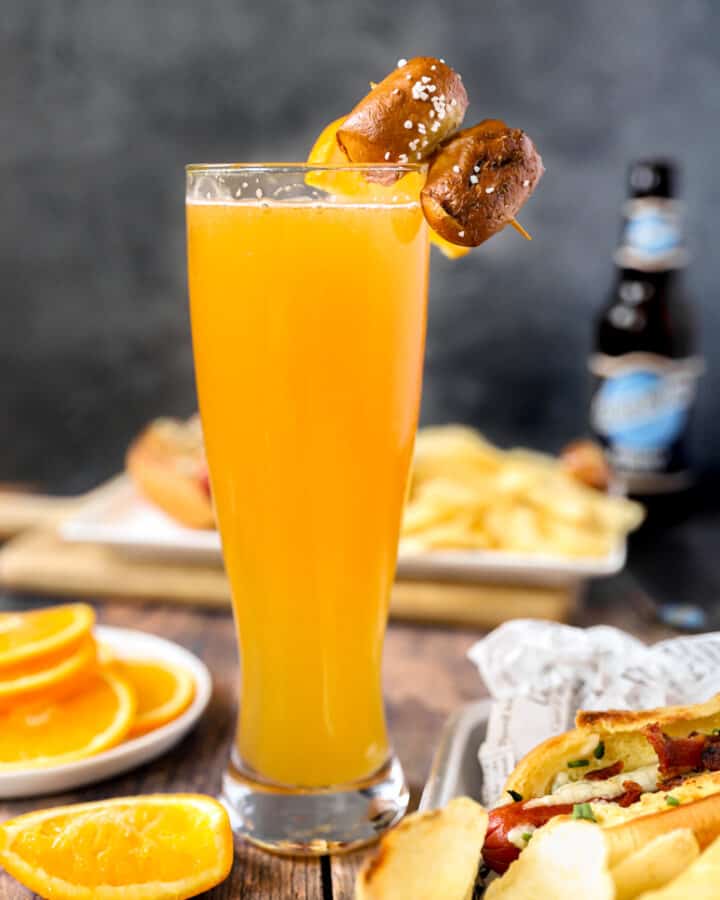 a beermosa on a wood surface with oranges and hotdog with chips