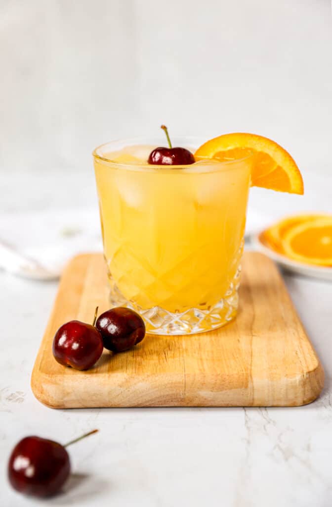 the cocktail on a wood cutting board with cherries in the foreground and a plate of orange slices in the background.