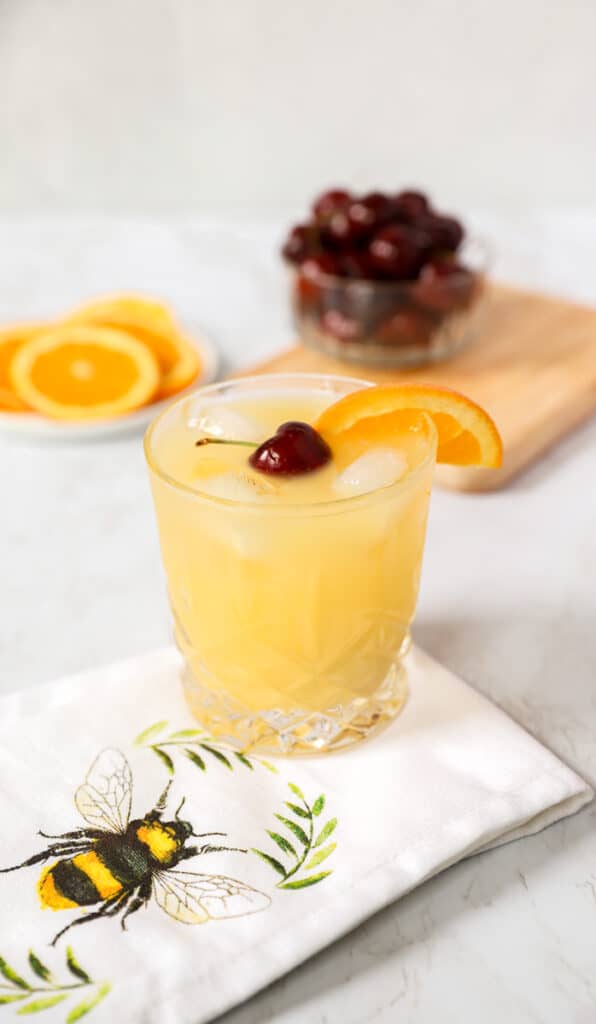 black eyed Susan cocktail on a linen napkin with an embroidered bee. Cherries and orange slices in the back.