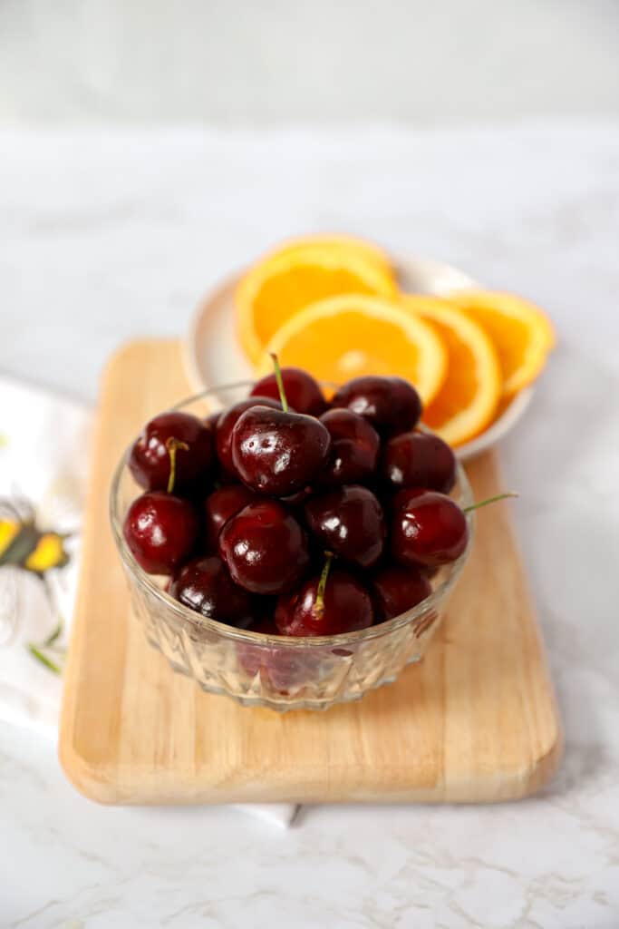 a bowl of fresh cherries and a plate of orange slices on a wood cutting board.