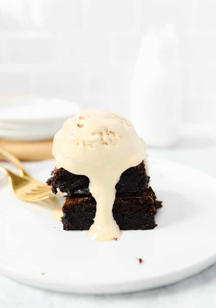 two stacked brownies smothered in coffee ice cream on a round white plate with two gold forks.