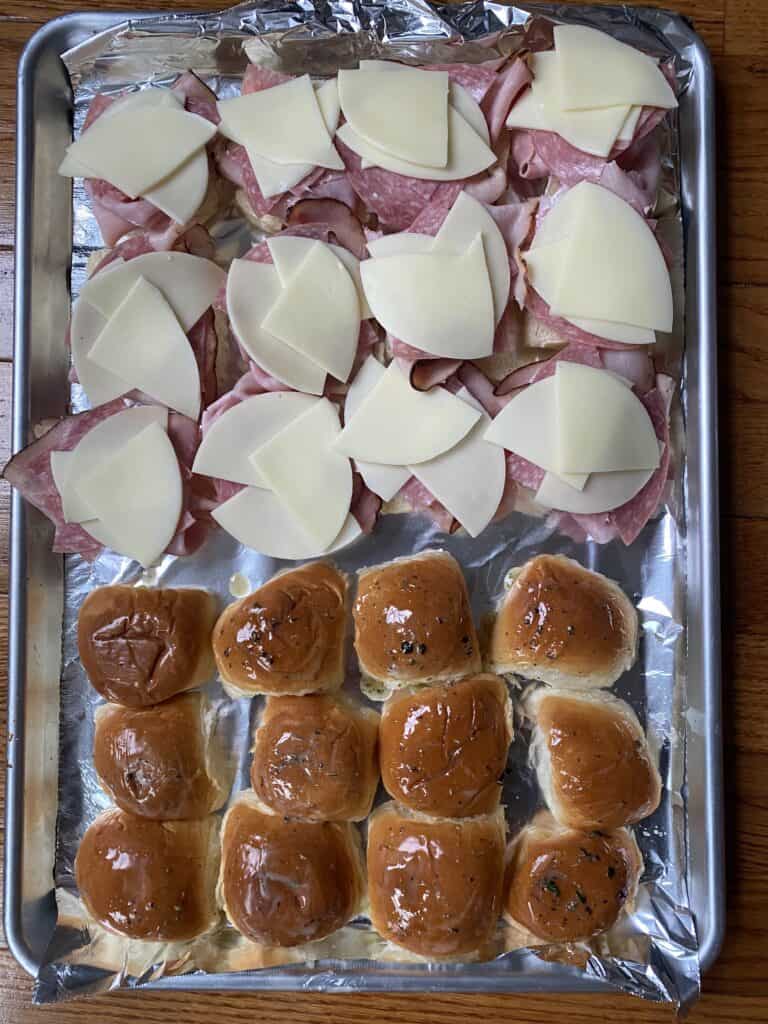 overheat shot of the bottom buns layered with the cheeses and meats and the top buns brushed with the herbed butter on a foil lined baking sheet.