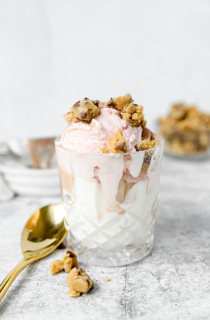 strawberry ice cream in a glass topped with the cookie dough pieces and a gold spoon.