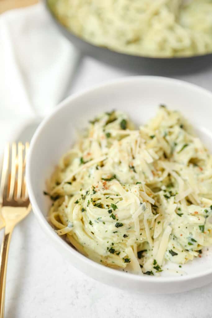 angled shot of the basil Alfredo swirled pasta noodles in a white bowl, with gold forks to the left. A pan of the pasta blurred in the background.