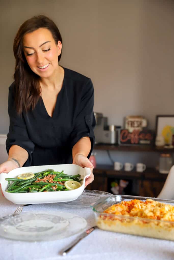 Catherine standing behind a white tablecloth covered table placing down a white casserole dish with green beans and lemons. On the table are clear glass plates, a casserole dish of mac and cheese and silver forks.