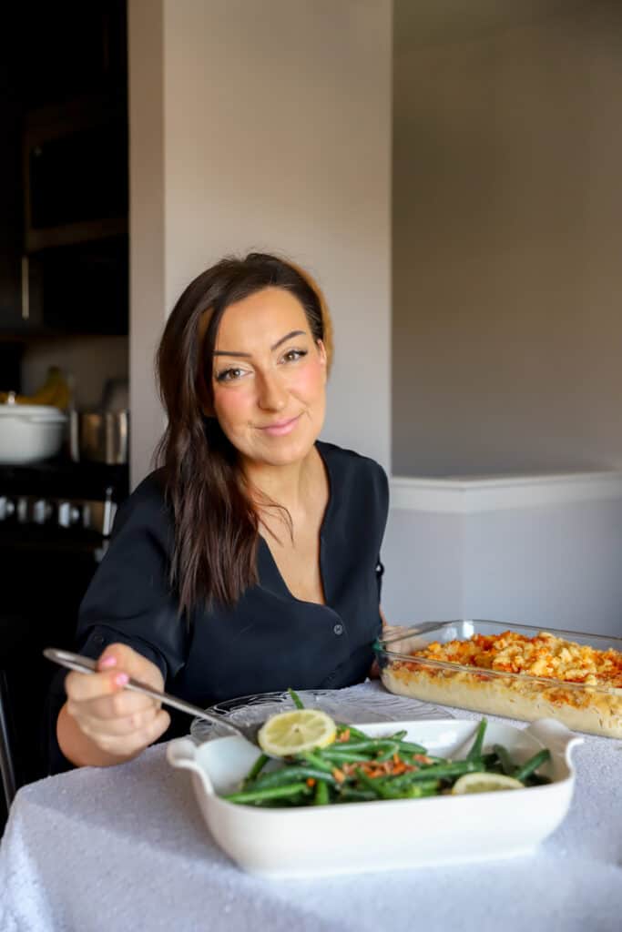 Catherine in a black dress sitting at a table serving a green bean casserole with lemon slices. A casserole of mac and cheese in front of her on a white tablecloth table.