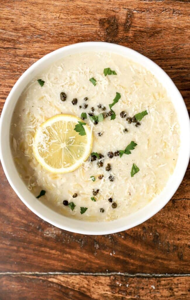 overhead photo of the chicken orzo lemon soup topped with fried capers, parsley, lemon slice in a white bowl on a wood surface.