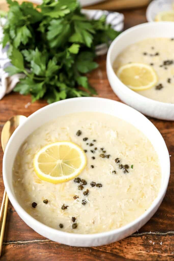 45 degree angled photo of the soup in a white bowl, topped with fried capers and a lemon slice A gold spoon sits to the left over the bowl on a wood surface.