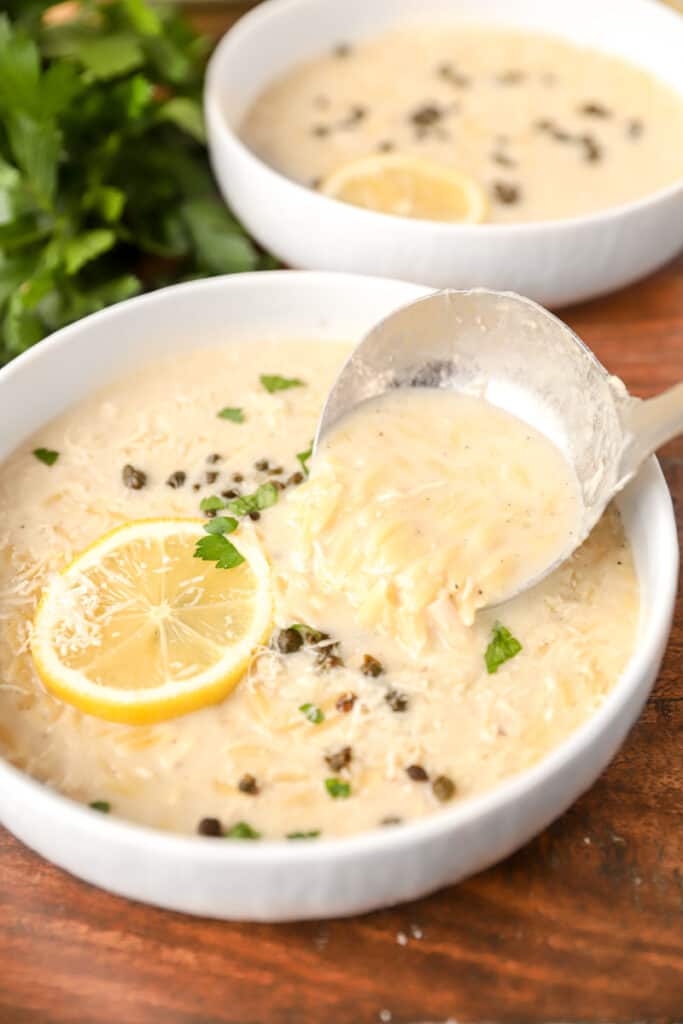 a silver ladle pouring the orzo chicken soup into a white bowl filled with the soup, topped with a lemon slice and fried capers. The bowl is on a warm wood surface.