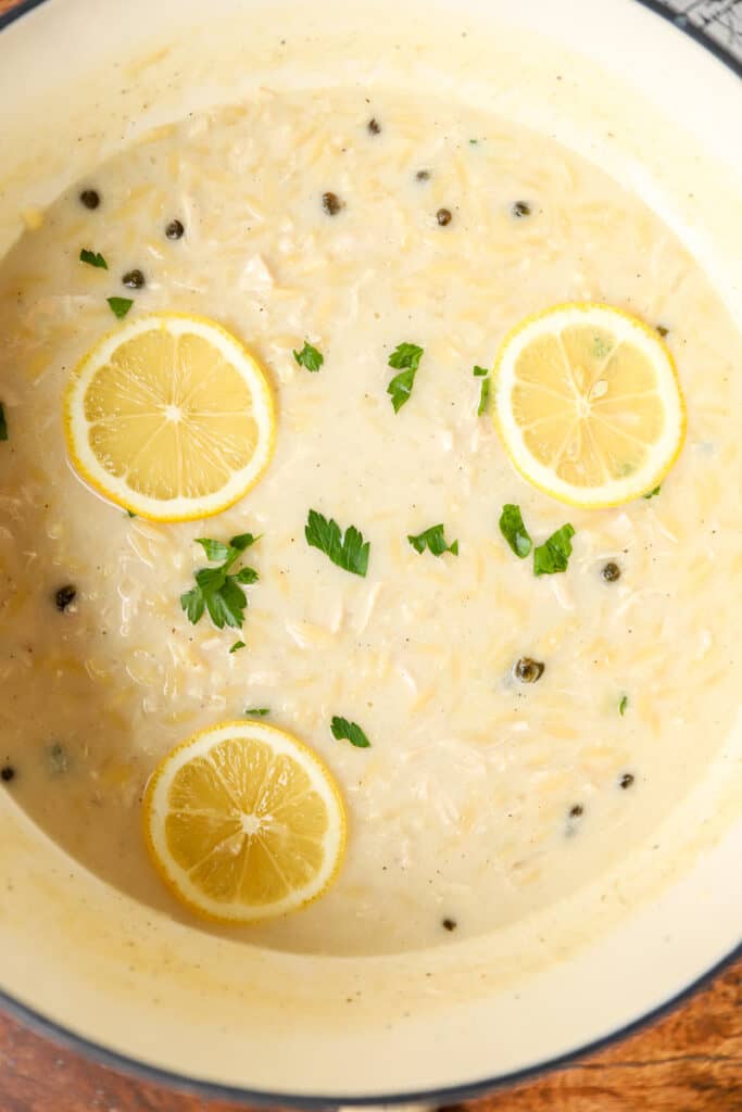 overhead photo of the creamy lemon chicken orzo soup in a Dutch oven topped with parsley and lemon slices.