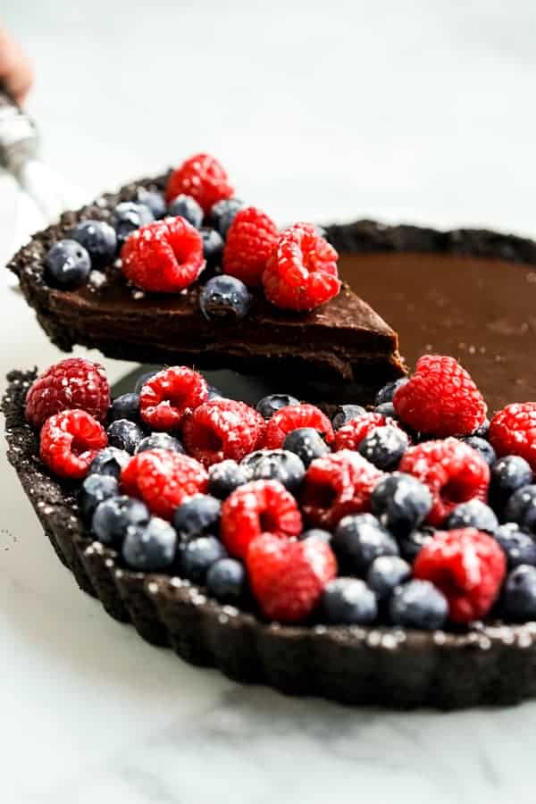 a chocolate tarte topped with raspberries and blueberries with a slice being removed by a pie server.