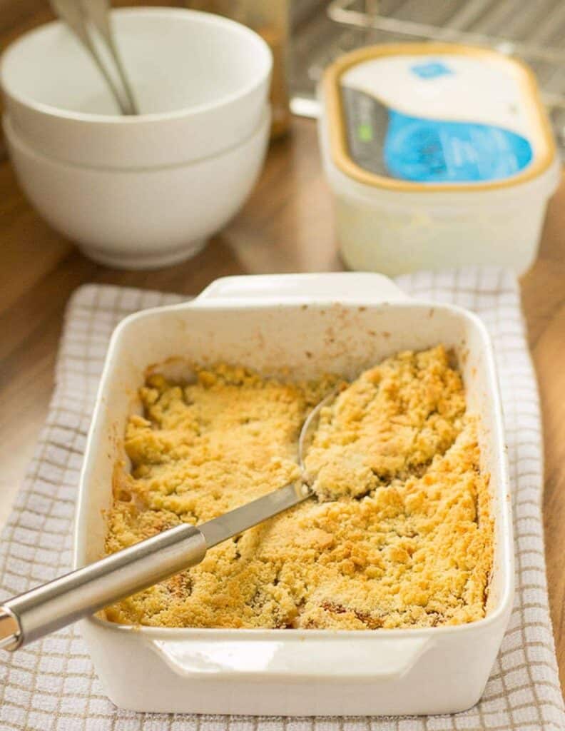 a white casserole dish with the cinnamon apple crumble and a large silver spoon, sitting on a white checkered pattern dish towel.