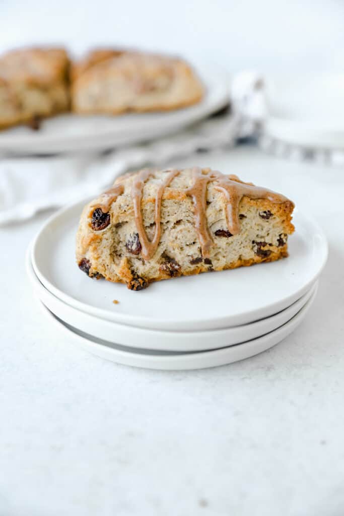 one large cinnamon raisin scone on a stack of 3 small white round plates. The larger plate of scones blurred in the back.