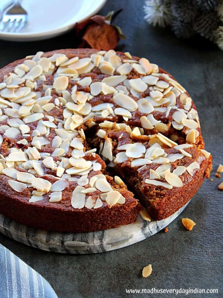 almond cake with a slice cut, plate is sitting on a dark grey surface and a plate and fork in the back.