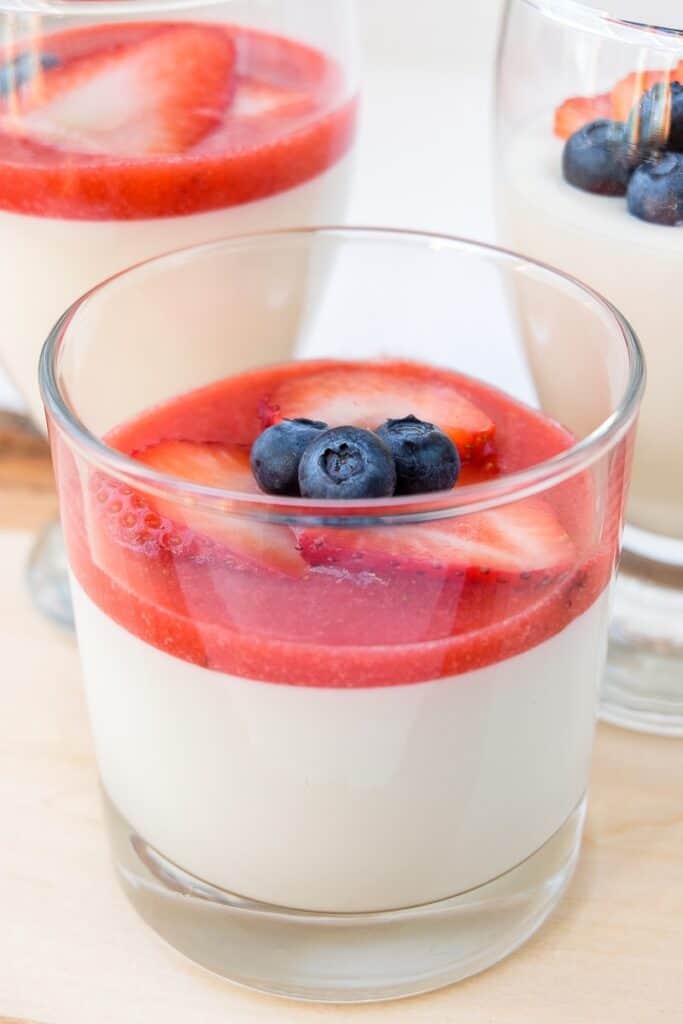 a serving of panna cotta in a clear glass. Topped with strawberries and blueberries.