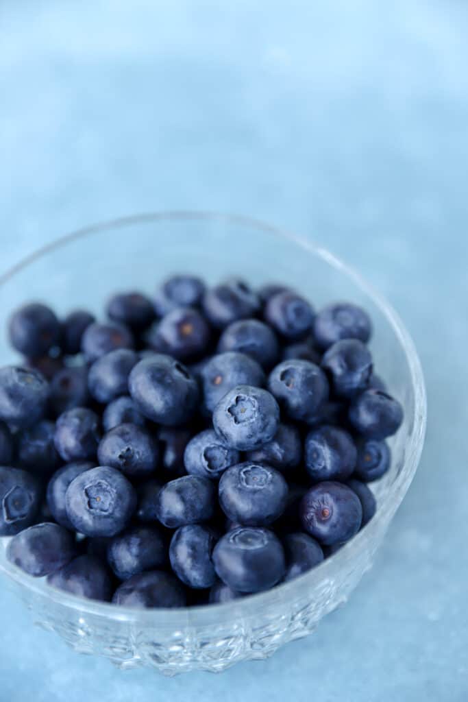 a clear bowl of fresh blueberries on a blue surface.