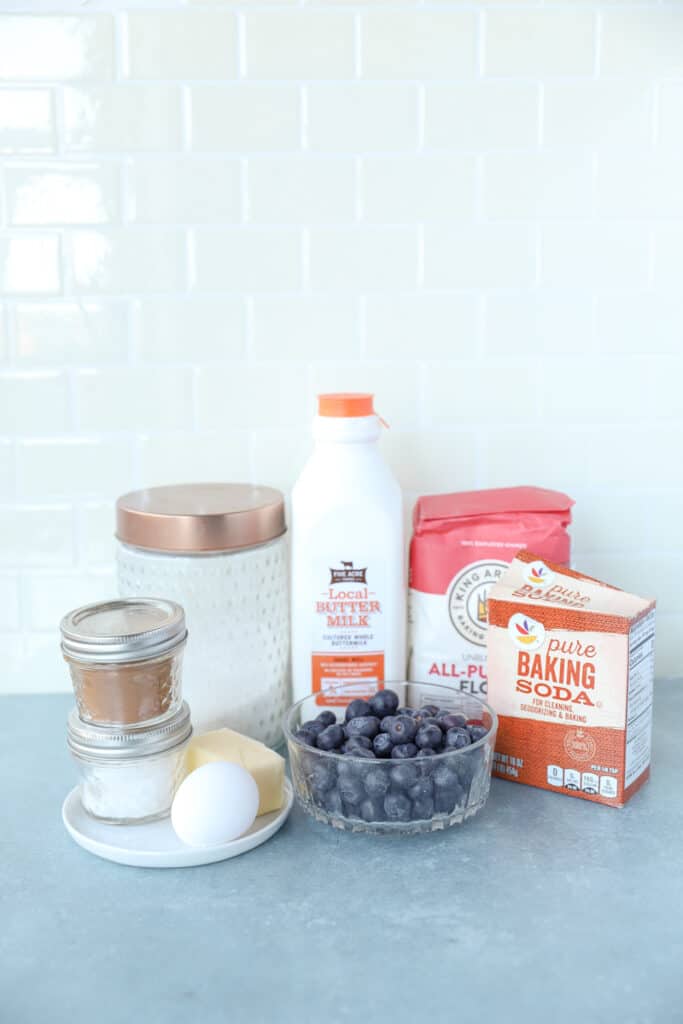 photo of the ingredients. containers of cinnamon, salt, and sugar. Butter and egg , bottle of buttermilk, bowl of blueberries, container of baking soda and bag of flour.