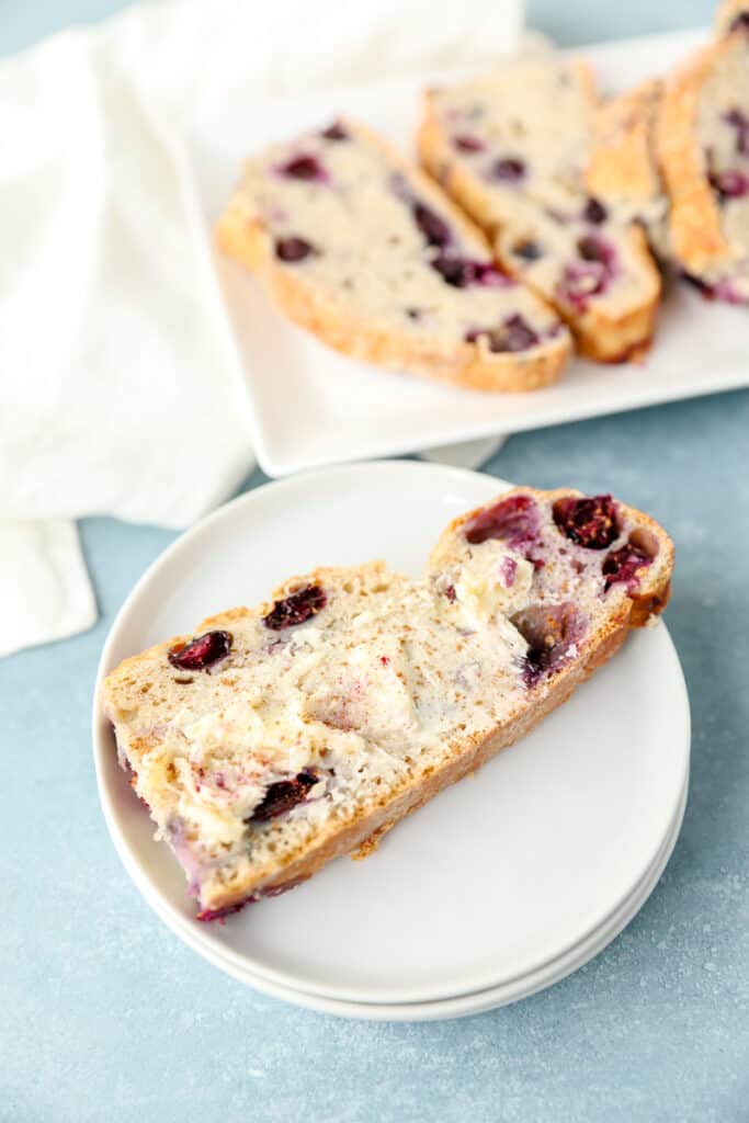 a buttered slice of blueberry soda bread on a stack of round white plates. In the background a rectangle white platter with more slices of the bread.