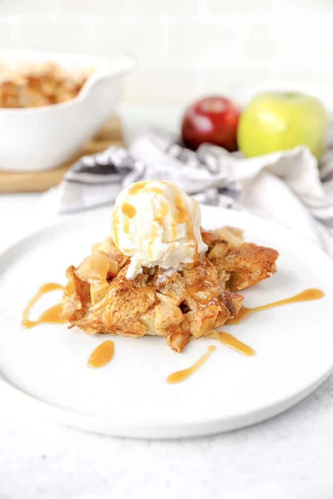 the biscuit bread pudding on a white plate with ice cream on top, red and green apples in the background.